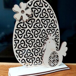 K&ouml;nnte beinhalten: Eine h&ouml;lzerne Ostereier-Dekoration mit einem keltischen Knotenmuster, einer kleinen Blume und einem Huhn, das ein Ei h&auml;lt. Das Ei steht auf einem Holzsockel.