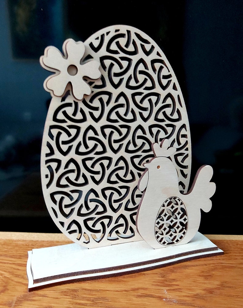 K&ouml;nnte beinhalten: Eine h&ouml;lzerne Ostereier-Dekoration mit einem keltischen Knotenmuster, einer kleinen Blume und einem Huhn, das ein Ei h&auml;lt. Das Ei steht auf einem Holzsockel.