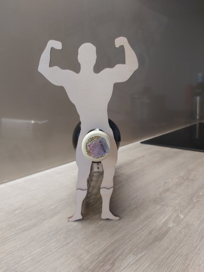 May include: Wooden wine bottle holder shaped like a person with arms raised in a strong pose. The bottle holder is made of light brown wood and is designed to hold a wine bottle.