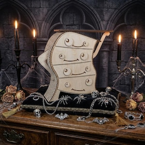 May include: A whimsical wooden dresser with a face design, set on a black velvet pillow with spider and skull accents. The scene includes lit candles, dried roses, and decorative jewelry, creating a spooky, gothic theme.