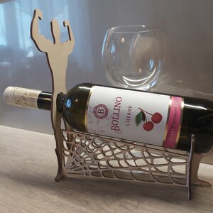 May include: Wooden wine bottle holder shaped like a muscular figure with a spiderweb design. The holder is holding a bottle of Bollino Cherry wine.