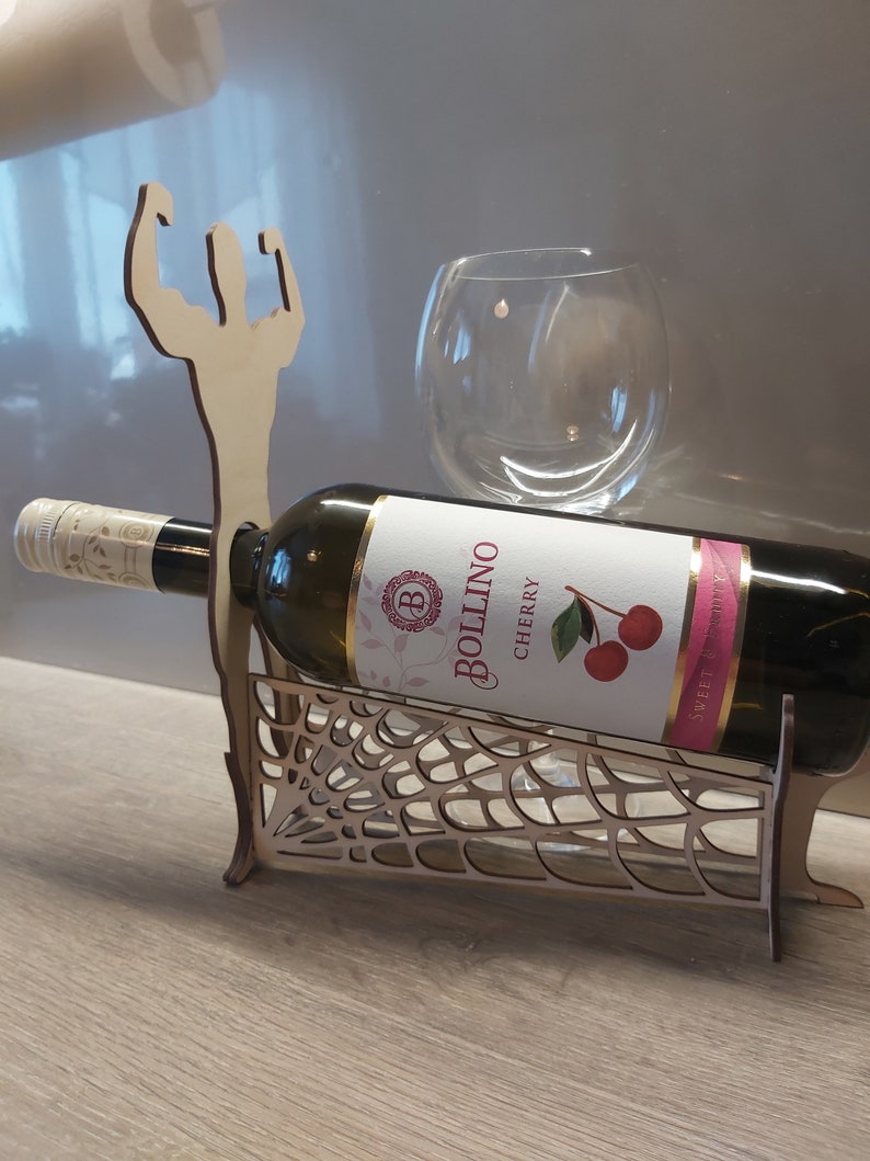 May include: Wooden wine bottle holder shaped like a muscular figure with a spiderweb design. The holder is holding a bottle of Bollino Cherry wine.