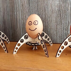 Puede incluir: Tres huevos marrones con caras dibujadas se muestran en soportes para huevos de madera con forma de cohetes. Los soportes para huevos tienen una base con forma de engranaje y est&aacute;n hechos de madera marr&oacute;n claro.