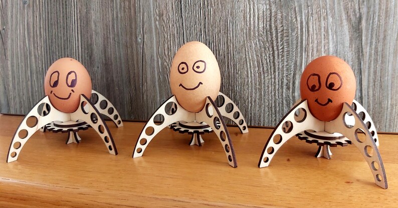Puede incluir: Tres huevos marrones con caras dibujadas se muestran en soportes para huevos de madera con forma de cohetes. Los soportes para huevos tienen una base con forma de engranaje y est&aacute;n hechos de madera marr&oacute;n claro.