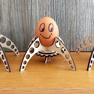 Puede incluir: Tres soportes para huevos de madera con forma de ara&ntilde;a con un huevo marr&oacute;n con una cara sonriente en el soporte central.