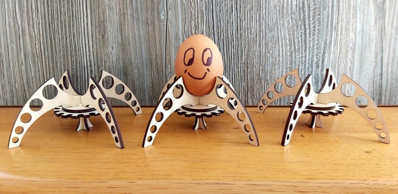 Puede incluir: Tres soportes para huevos de madera con forma de ara&ntilde;a con un huevo marr&oacute;n con una cara sonriente en el soporte central.