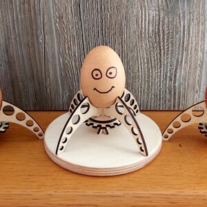 Puede incluir: Tres soportes para huevos de madera con huevos marrones que tienen caras dibujadas. Los soportes para huevos tienen forma de tr&iacute;pode con un engranaje en el centro. Los soportes para huevos est&aacute;n sobre una superficie de madera.