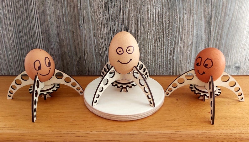 Puede incluir: Tres soportes para huevos de madera con huevos marrones que tienen caras dibujadas. Los soportes para huevos tienen forma de tr&iacute;pode con un engranaje en el centro. Los soportes para huevos est&aacute;n sobre una superficie de madera.
