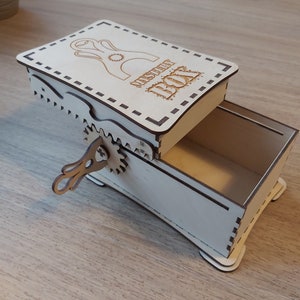 May include: A wooden box with a hinged lid and a small, intricate metal crank on the side. The lid is decorated with a laser-cut design and the words "Mystery Box".