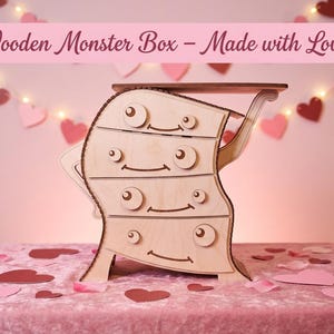 May include: A wooden monster box with four drawers, featuring a whimsical design with eyes and smiles. The box is made of light-colored wood and has a small shelf on top. The text "Wooden Monster Box - Made with Love!" is displayed above the box.