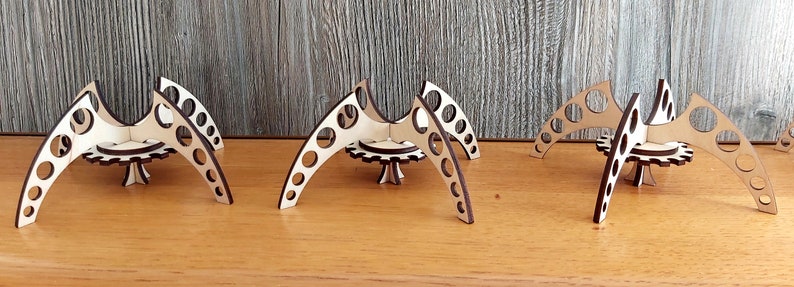 Puede incluir: Tres soportes geom&eacute;tricos de madera cortados con l&aacute;ser, con una base circular y tres patas curvas. Cada soporte tiene un patr&oacute;n circular de agujeros en las patas.