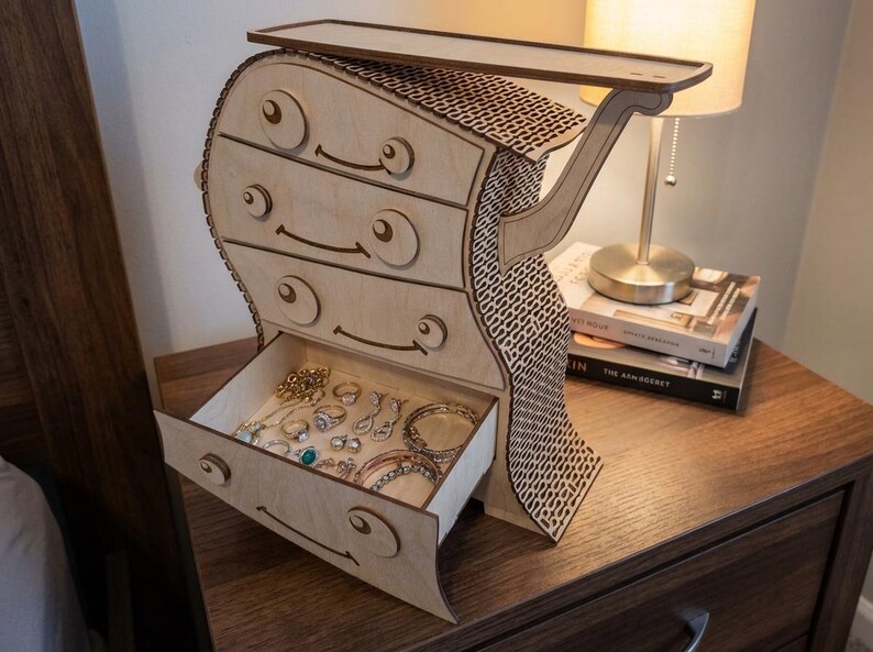 May include: A whimsical wooden jewelry chest with four drawers and a top shelf. The chest is light-colored with a unique design, featuring curved lines and decorative cutouts. The open drawer reveals various jewelry pieces.
