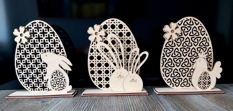 K&ouml;nnte beinhalten: Drei h&ouml;lzerne Ostereier-Dekorationen mit verschiedenen Designs. Das erste Ei hat ein geometrisches Muster und ein Kaninchen. Das zweite Ei hat ein gewebtes Muster und zwei Kaninchen. Das dritte Ei hat ein keltisches Knotenmuster und ein K&uuml;ken.