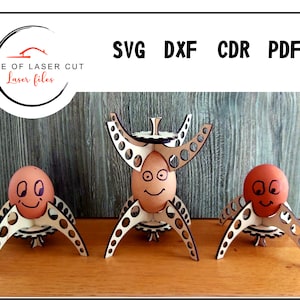 Puede incluir: Tres soportes para huevos de madera con forma de cohete con una cara sonriente dibujada en cada huevo. Los soportes para huevos est&aacute;n sobre una superficie de madera. SVG DXF CDR PDF.