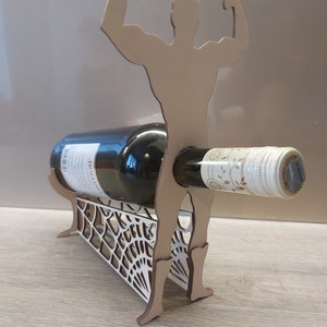 May include: Wooden wine bottle holder shaped like a muscular figure with arms raised. The figure is holding a bottle of wine in its arms. The base of the holder is a decorative lattice design.