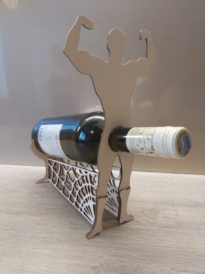 May include: Wooden wine bottle holder shaped like a muscular figure with arms raised. The figure is holding a bottle of wine in its arms. The base of the holder is a decorative lattice design.