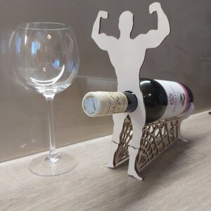 May include: White wooden wine bottle holder shaped like a person lifting weights. The holder has a bottle of wine in it and a wine glass is on the table next to it.
