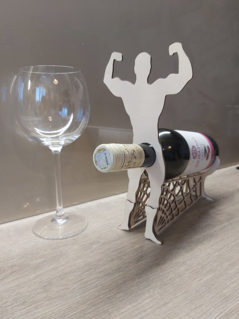 May include: White wooden wine bottle holder shaped like a person lifting weights. The holder has a bottle of wine in it and a wine glass is on the table next to it.