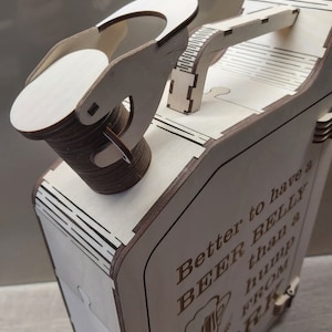 Puede incluir: Caja de madera con tapa con bisagras y asa. La caja tiene forma de bid&oacute;n de gasolina y tiene el texto "Mejor tener una BARRIGA DE CERVEZA que una joroba del TRABAJO" grabado en el lateral.