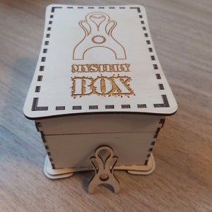 May include: A white wooden box with a decorative design on the lid. The lid has a black and white dashed border and the text "MYSTERY BOX" is engraved on the lid. The box has a small wooden stand with a decorative design.
