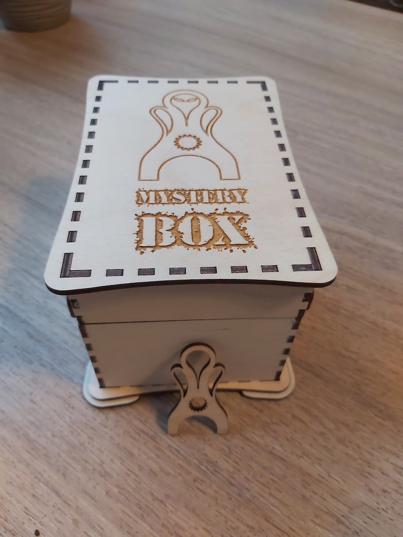 May include: A white wooden box with a decorative design on the lid. The lid has a black and white dashed border and the text "MYSTERY BOX" is engraved on the lid. The box has a small wooden stand with a decorative design.