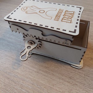 May include: A wooden box with a hinged lid and a small, intricate gear mechanism. The lid is engraved with the words "Mystery Box".