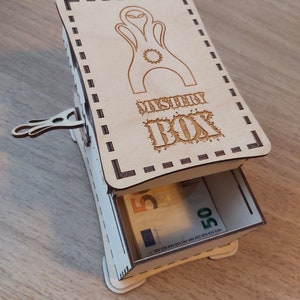 May include: A wooden box with a drawer that opens to reveal money. The box is decorated with a laser-cut design of an alien and the words "Mystery Box".
