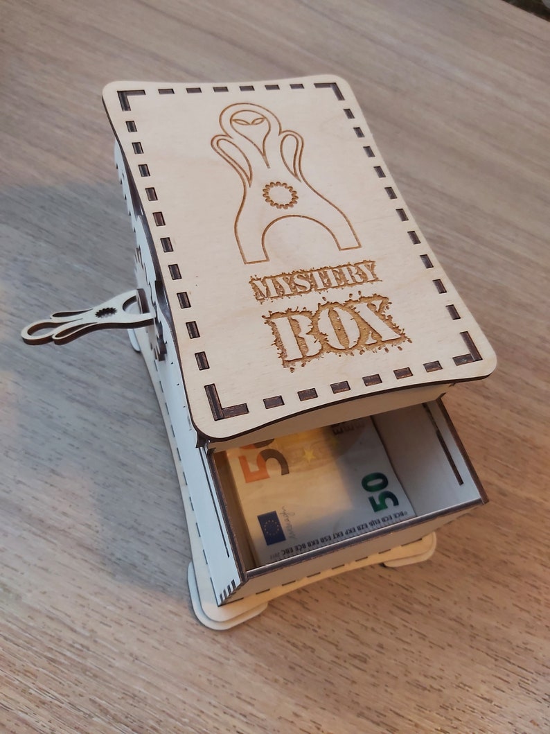 May include: A wooden box with a drawer that opens to reveal money. The box is decorated with a laser-cut design of an alien and the words "Mystery Box".