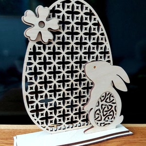 K&ouml;nnte beinhalten: Eine h&ouml;lzerne Osterhasenfigur mit einem Ausschnittmuster. Der Hase h&auml;lt ein dekoratives Ei mit einem geometrischen Muster. Der Hase steht vor einem gro&szlig;en Ei mit einem geometrischen Muster. Das Ei ist mit einer Blume verziert.