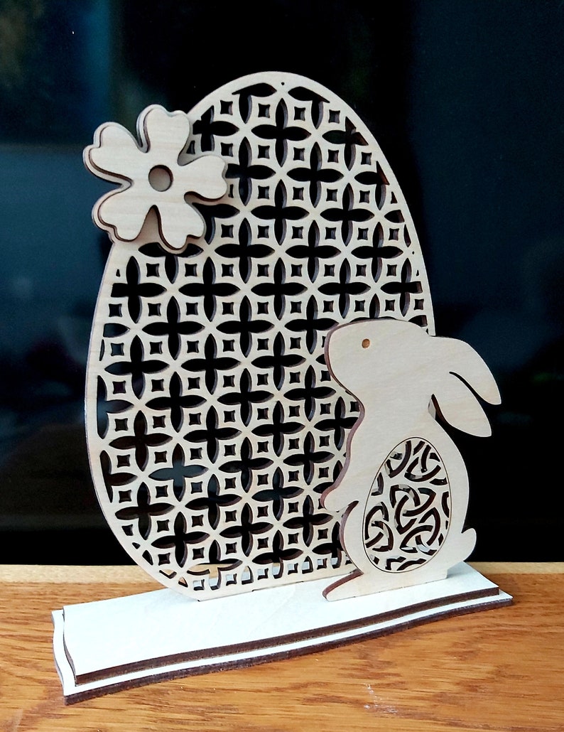 K&ouml;nnte beinhalten: Eine h&ouml;lzerne Osterhasenfigur mit einem Ausschnittmuster. Der Hase h&auml;lt ein dekoratives Ei mit einem geometrischen Muster. Der Hase steht vor einem gro&szlig;en Ei mit einem geometrischen Muster. Das Ei ist mit einer Blume verziert.