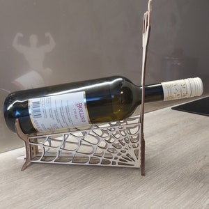 May include: Wooden wine bottle holder shaped like a spider web. The holder is made of light brown wood and has a bottle of Bollino Chianti wine resting in it.
