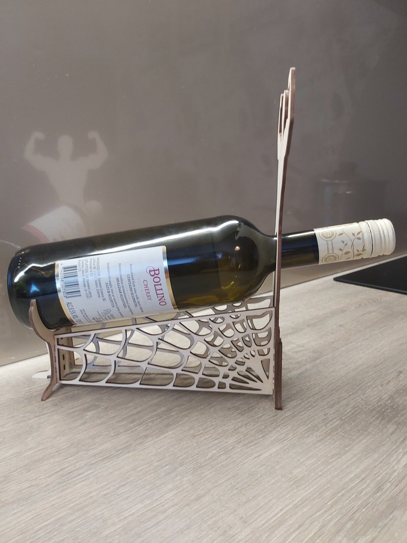 May include: Wooden wine bottle holder shaped like a spider web. The holder is made of light brown wood and has a bottle of Bollino Chianti wine resting in it.