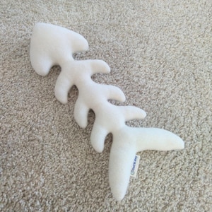 Può includere: Giocattolo di peluche bianco a forma di lisca di pesce con un'etichetta "Cozy Joy" attaccata.
