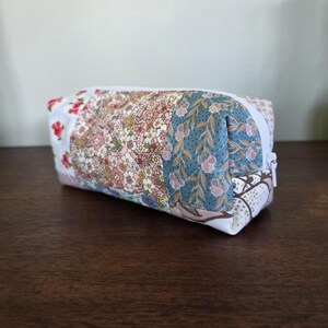 May include: A rectangular pencil case with a white zip. The case is made of patchwork fabric with floral patterns in pink, blue, and white. The case is on a dark wooden surface.