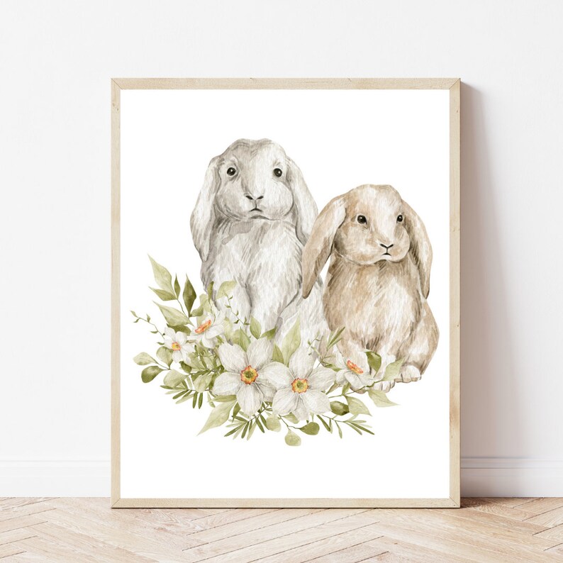 K&ouml;nnte beinhalten: Ein Aquarellbild von zwei Kaninchen, die in einem Bett aus wei&szlig;en Blumen sitzen. Das Kaninchen links ist wei&szlig; mit grauen Markierungen, und das Kaninchen rechts ist braun. Die Blumen sind von gr&uuml;nen Bl&auml;ttern umgeben.