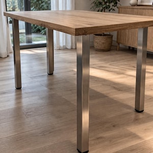 Op de afbeelding: Een rechthoekige eettafel met een lichtbruin houten tafelblad met zichtbare houtnerf en vier stevige, vierkante poten van geborsteld roestvrij staal. Elke poot is aan de onderkant voorzien van een kleine zwarte beschermdop.