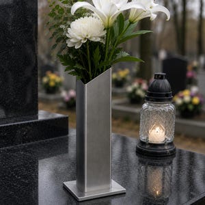 Könnte beinhalten: Eine hohe, rechteckige Vase aus gebürstetem Metall mit schräg verlaufender Oberkante, gefüllt mit einem Strauß aus weißen Lilien und einer weißen Chrysantheme. Die Vase steht auf einem quadratischen Sockel auf einer polierten schwarzen Granitplatte.