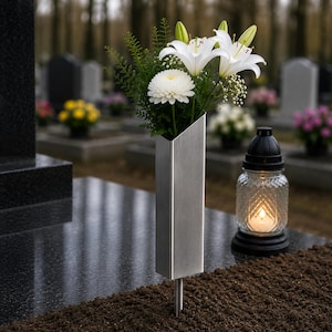 Może przedstawiać: Wazon nagrobny ze szczotkowanego metalu ze skośnym otworem, zawierający bukiet białych lilii, białą chryzantemę i zielone liście. Szklany znicz z zapaloną świecą stoi na ciemnej ziemi obok wazonu.