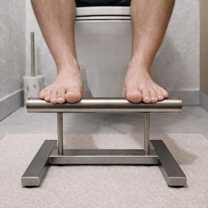 May include: A stainless steel foot stool with a horizontal bar, designed to assist with bathroom use. The stool is placed on a light-colored rug in front of a toilet. The person's feet are resting on the bar.