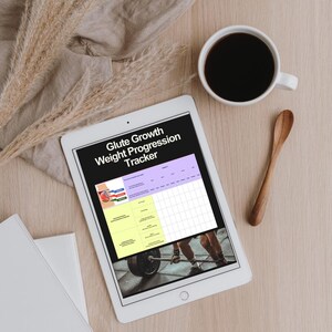 May include: A white tablet displaying a "Glute Growth Weight Progression Tracker" with a workout graphic. A cup of coffee and a wooden spoon sit nearby on a light wood surface. Dried ornamental grass and a folded cloth are also present.