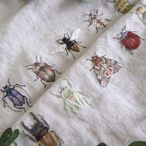 Puede incluir: Primer plano de una tela de lino con bordados de insectos. La tela muestra una abeja, escarabajos y una polilla, todos bordados con detalle. Los insectos son de varios colores, incluyendo rojo, marrón, amarillo y azul. Hay hojas verdes en las esquinas.