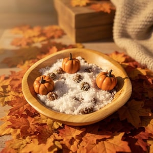 May include: A round, light-colored wooden bowl filled with white granular material, two small orange pumpkin figurines, and several small brown textured spheres, arranged on a surface covered with scattered orange and yellow autumn leaves.