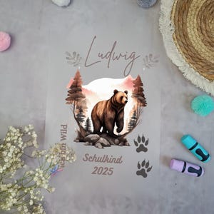 Könnte beinhalten: Ein graues Poster mit einer Illustration eines braunen Bären in einer Waldszene. Der Name "Ludwig" steht über dem Bären, darunter "Schulkind 2025". Pfotenabdrücke und der Text "Ich bin Wild" sind ebenfalls vorhanden.