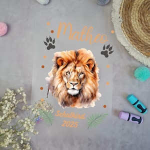 Könnte beinhalten: Eine transparente Folie mit einer Aquarellillustration eines Löwenkopfes mit goldener Mähne. Der Name "Matheo" steht in orangefarbener Schrift über dem Löwen, mit Pfotenabdrücken und Punkten. Der Text "Schulkind 2025" steht darunter.