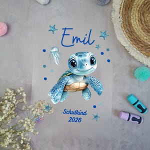 Könnte beinhalten: Eine transparente Folie mit einer Cartoon-Schildkröte in Blau und Braun, dem Namen "Emil" in blauer Schrift und dem Text "Schulkind 2026". Das Design umfasst blaue Sterne, Punkte und Quallen.