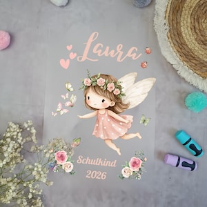 Könnte beinhalten: Eine transparente Folie mit einer Cartoon-Fee in einem rosa Kleid, Blumenkranz und Flügeln. Der Name "Laura" steht über der Fee, darunter "Schulkind 2026". Rosa Schmetterlinge und Blumen schmücken das Design, ideal für Kinderzimmer.