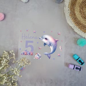 Könnte beinhalten: Ein klares, durchsichtiges Blatt mit einem lila Delfin, einer lila Zahl 5 und dem Namen "Helena" in lila Schrift. Der Hintergrund hat einen helllila Farbton und zeigt kleine weiße Sterne, lila Punkte und eine lila Muschel.