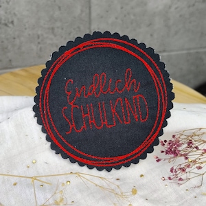 Könnte beinhalten: Ein schwarzer Filz-Kreis mit roter Stickerei und dem Text "Endlich Schulkind" in Rot.