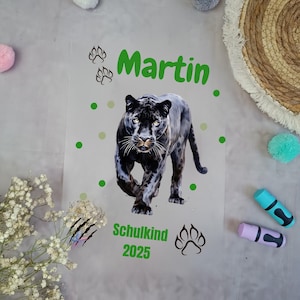 Könnte beinhalten: Eine transparente Folie mit einer Illustration eines schwarzen Panthers. Der Name "Martin" steht in grüner Schrift, zusammen mit den Worten "Schulkind 2025". Das Design umfasst Pfotenabdrücke und grüne Punkte.