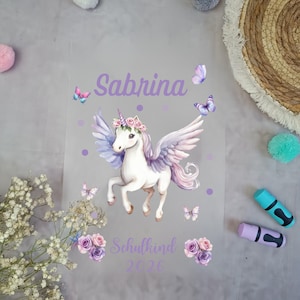 Könnte beinhalten: Eine transparente Folie mit einer Aquarell-Illustration eines weißen Einhorns mit lila und rosa Flügeln, einem Blumenkranz und dem Namen "Sabrina" in lila Schrift. Schmetterlinge und florale Akzente umgeben das Einhorn, mit dem Text "Schulkind 2026" darunter.